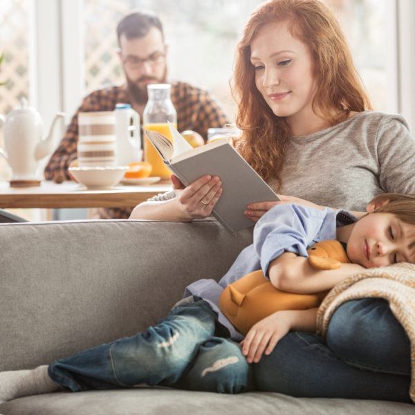 Sleeping son lying on mother's laps while she is reading a book and father eating in the background